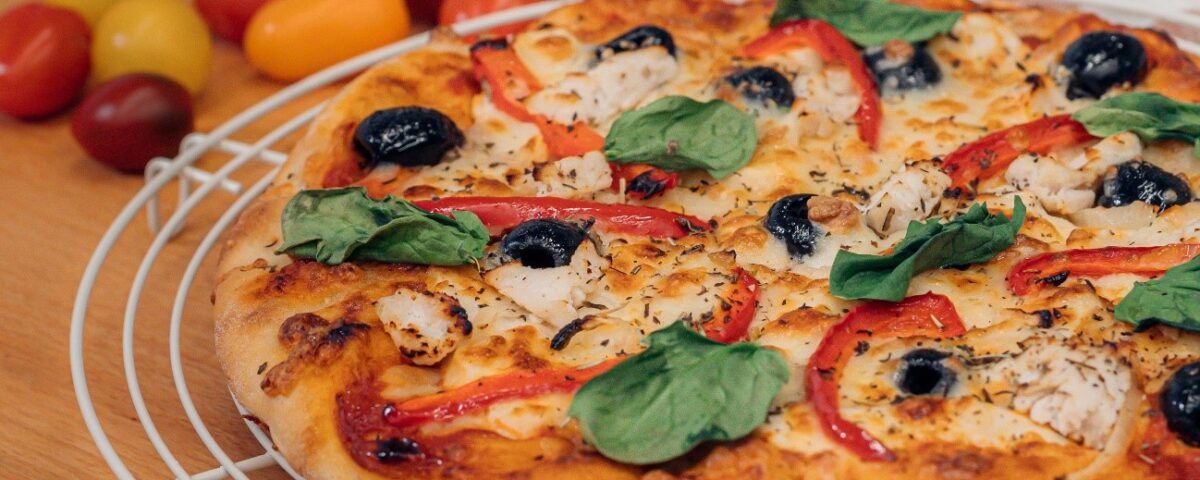 Una pizza recién horneada sobre una rejilla blanca, con pollo, aceitunas negras, tiras de pimiento rojo y hojas frescas de albahaca, con tomates cherry al fondo.