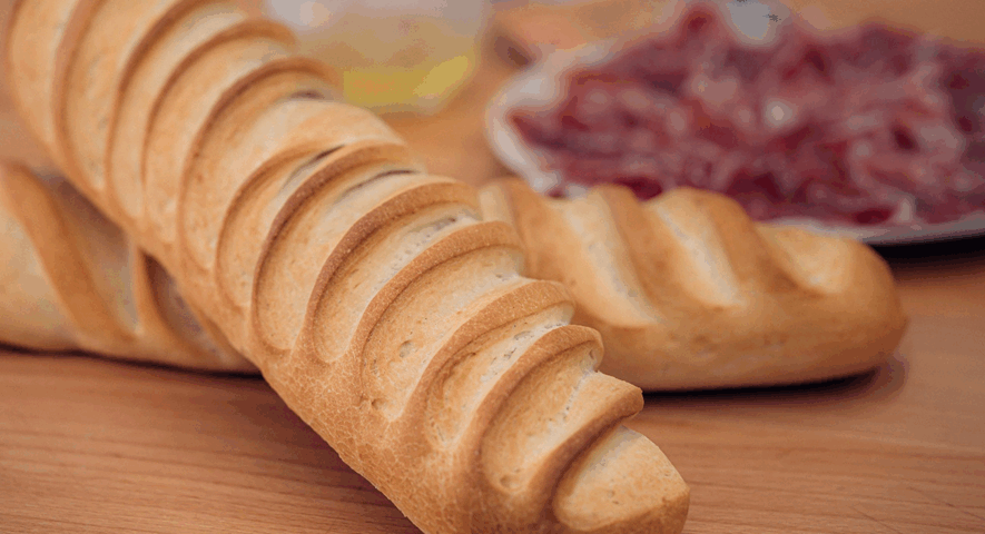 Dos barras de pan candeal con un greñado profundo y dorado sobre una mesa de madera, con un plato de jamón ibérico al fondo