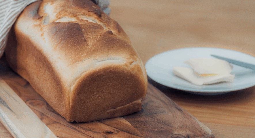 Una pieza entera de pan de molde dorado sobre una tabla de madera, envuelto parcialmente en un paño de cocina y acompañado de mantequilla en un plato