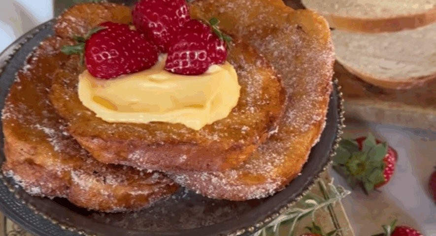Tres torrijas doradas y rebozadas en azúcar apiladas sobre un plato decorado, coronadas con una porción de crema y fresas maduras, con rebanadas de pan al fondo