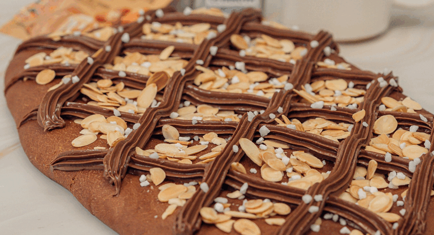 Coca de San Juan de masa oscura de chocolate, decorada con un enrejado de crema, almendras laminadas y azúcar perlado