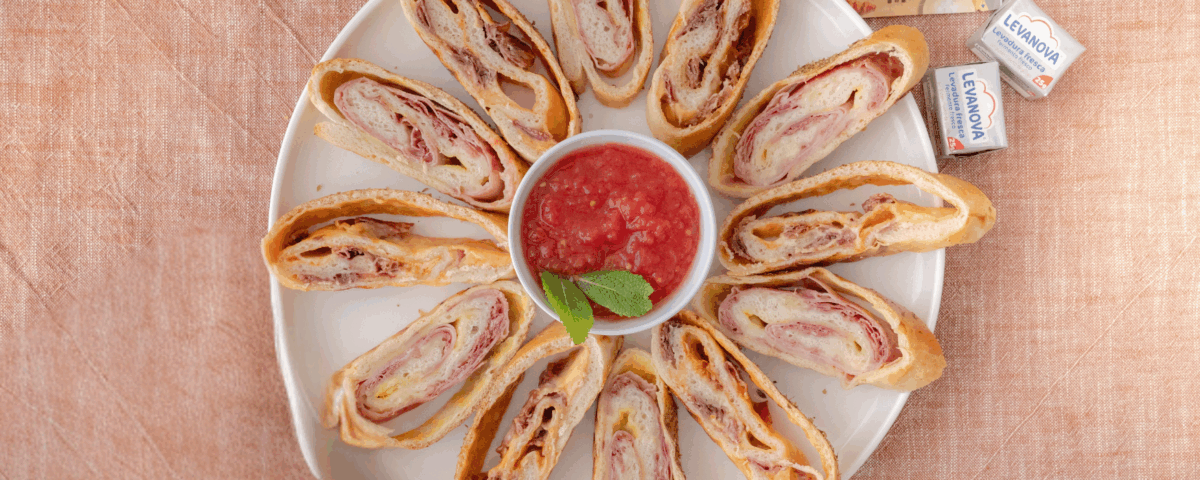 Rodajas de stromboli rellenas de jamón y queso dispuestas en círculo alrededor de un cuenco de salsa de tomate, con productos Levanova al lado