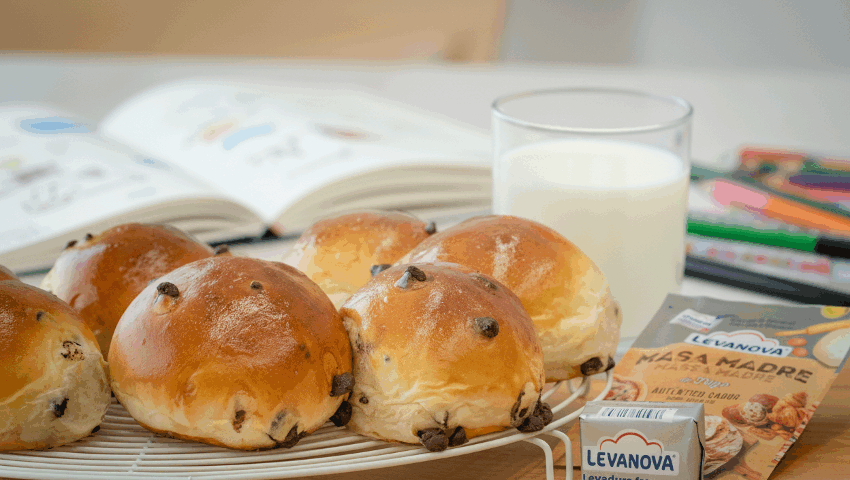 Varios Doowaps con pepitas de chocolate sobre una rejilla, junto a un vaso de leche, levadura Levanova y material escolar al fondo