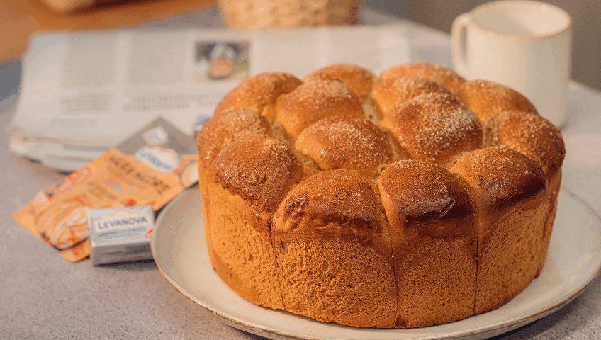 Un brioche alto y esponjoso formado por bollitos con azúcar por encima, presentado en un plato junto a una taza, un periódico y levadura Levanova