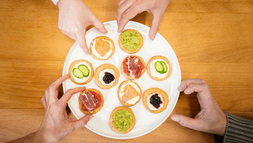 Vista aérea de varias manos cogiendo canapés de pan marinero con diferentes rellenos (salmón, jamón, aguacate y mermelada) dispuestos en un plato blanco sobre una mesa de madera