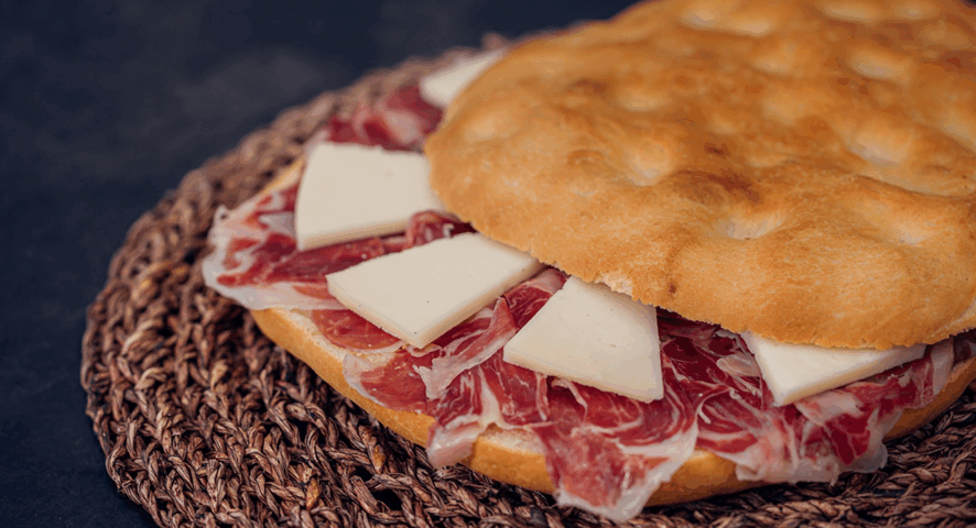 Torta de Aranda abierta y rellena generosamente con lonchas de jamón ibérico y cuñas de queso, presentada sobre un mantel individual de rafia