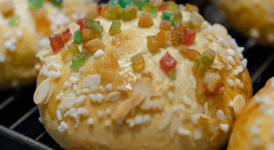 Primer plano de un mini roscón dorado y brillante, decorado con trozos de fruta escarchada de colores, azúcar perlado y láminas de almendra sobre una rejilla