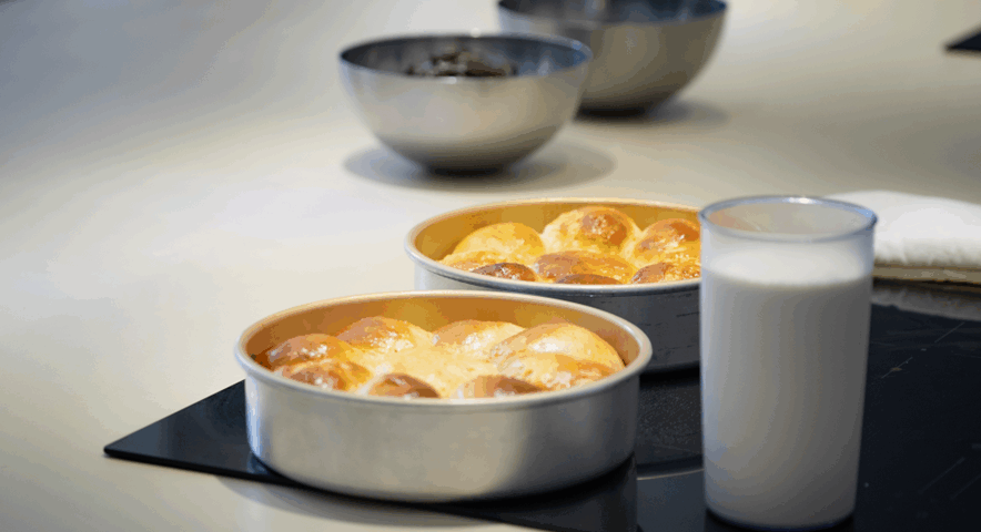 Dos moldes redondos con pan japonés dorado sobre una placa de inducción, acompañados de un vaso de leche y cuencos metálicos al fondo