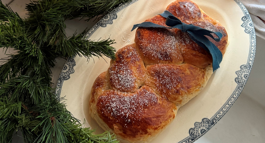 Brioche trenzado con azúcar glass y un lazo azul sobre una fuente clásica, decorado con ramas de pino navideñas