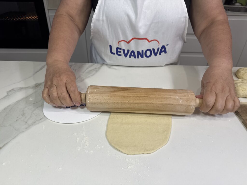 Primer plano de una persona con un delantal de Levanova estirando una porción de masa con un rodillo de madera sobre una encimera blanca