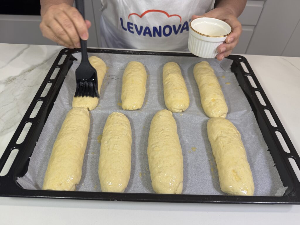 Persona pincelando con huevo batido varios bollos de masa dispuestos sobre una bandeja de horno con papel vegetal